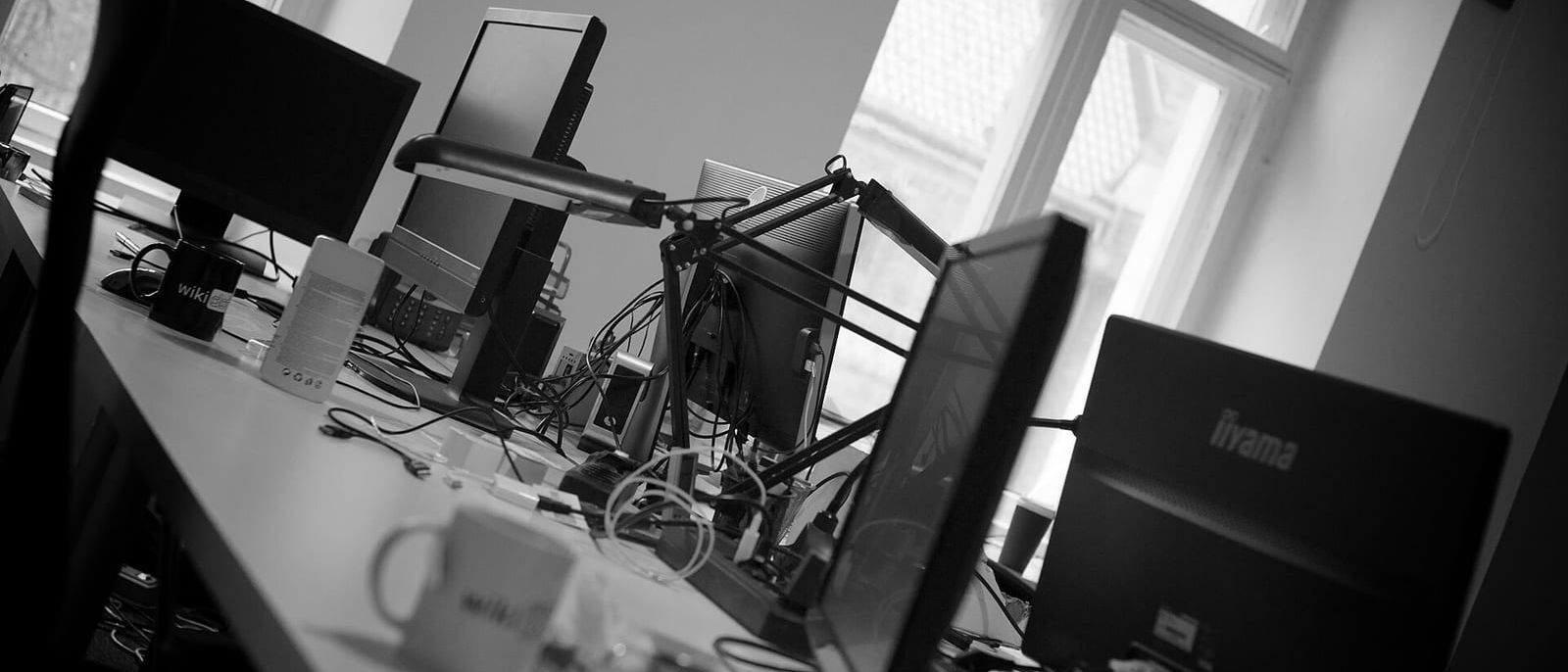 Black-and-white office with several monitors, cables and coffee mugs on a long desk