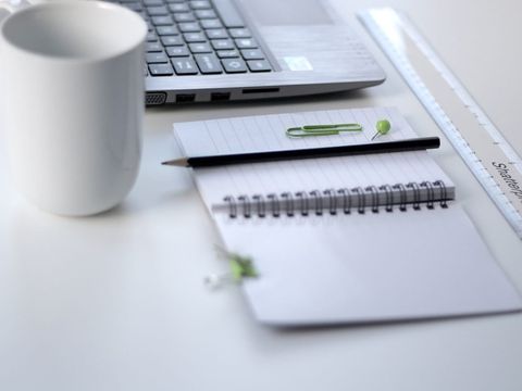 Laptop, white mug, spiral notepad with pencil and ruler on a bright desk