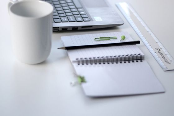 Laptop, white mug, spiral notepad with pencil and ruler on a bright desk