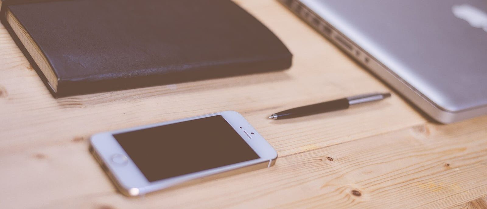 Smartphone, Kugelschreiber, Notizbuch und zugeklappter Laptop auf einem Holztisch