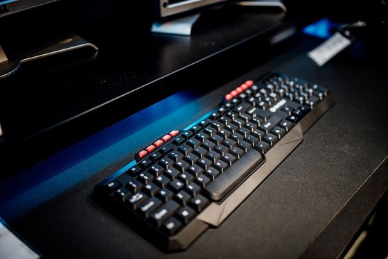 Schwarze Computertastatur auf dunklem Schreibtisch, dahinter Bildschirmfüße im blauen Licht