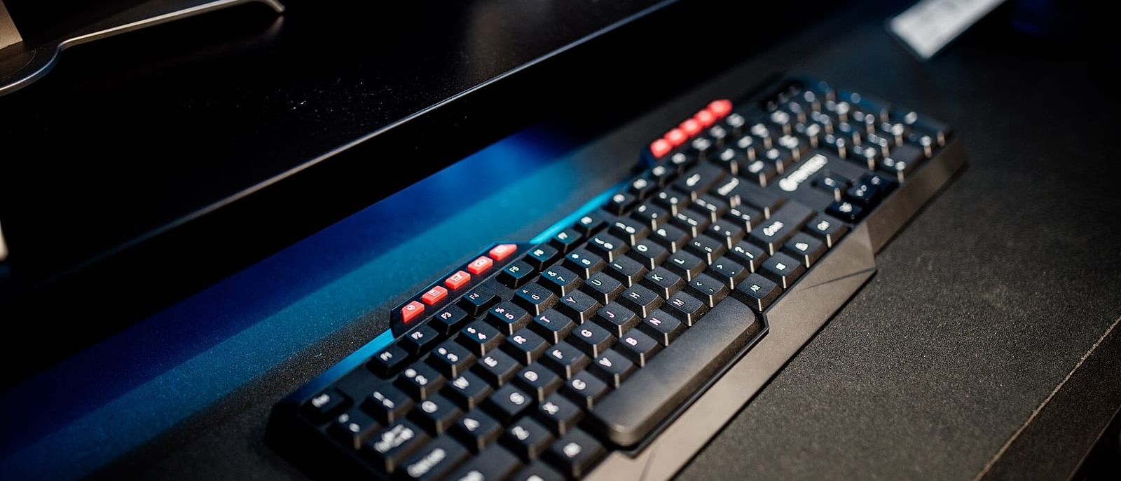 Black computer keyboard on a dark desk with monitor stands behind it in blue light